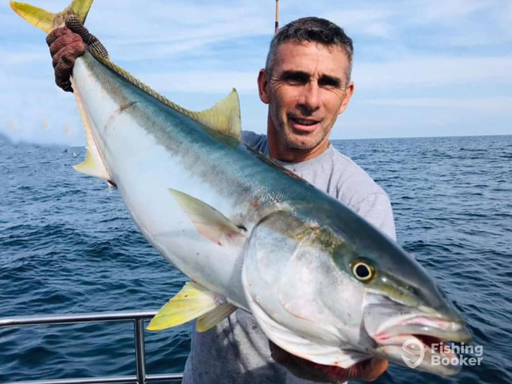 A man stands on a boat in the ocean on a sunny day holding a large Yellowtail Amberjack diagonally across his body