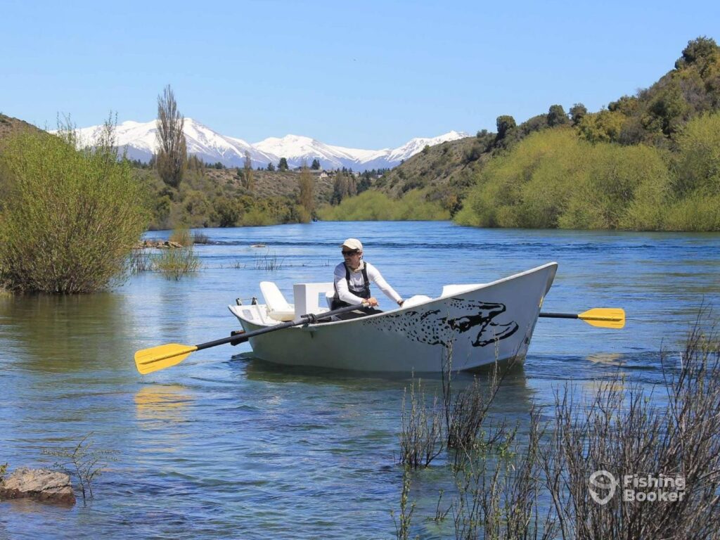 A fishing guide rows a small boat along a narrow river in Patagonia with snow-covered mountains in the background