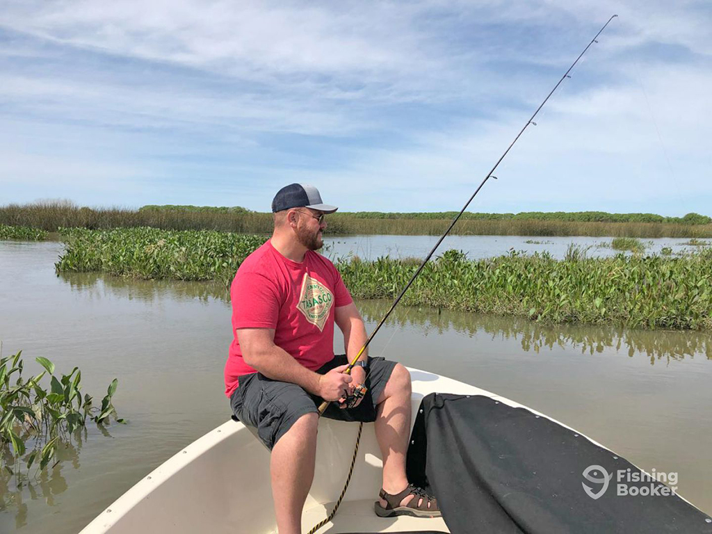 An angler sits at the front of a small boat in muddy, marshy waters while holding a long fishing rod on a sunny day