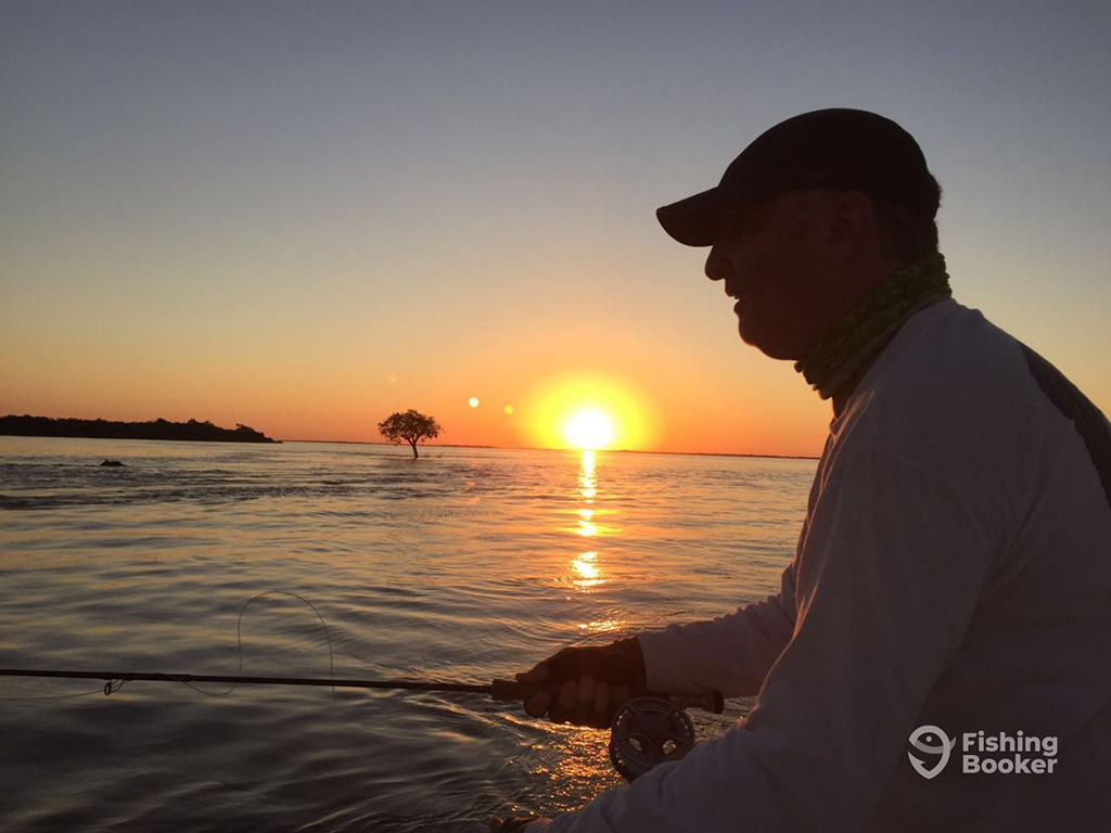 An angler in the shadow of the distant sun setting holds a fly fishing rod against a vast expanse of flooded water with a single tree in the distance 