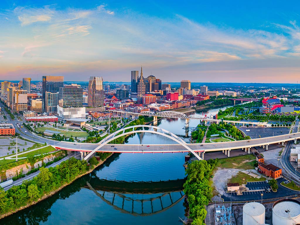 An aerial view looking down the Cumberland River towards the skyline of Nashville, TN, with the city's newest bridge in the foreground on a clear day