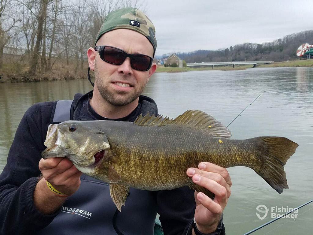 An angler in a back-to-front baseball cap and sunglasses holding Smallmouth Bass towards the camera on a cloudy day with the waters of a river visible behind him