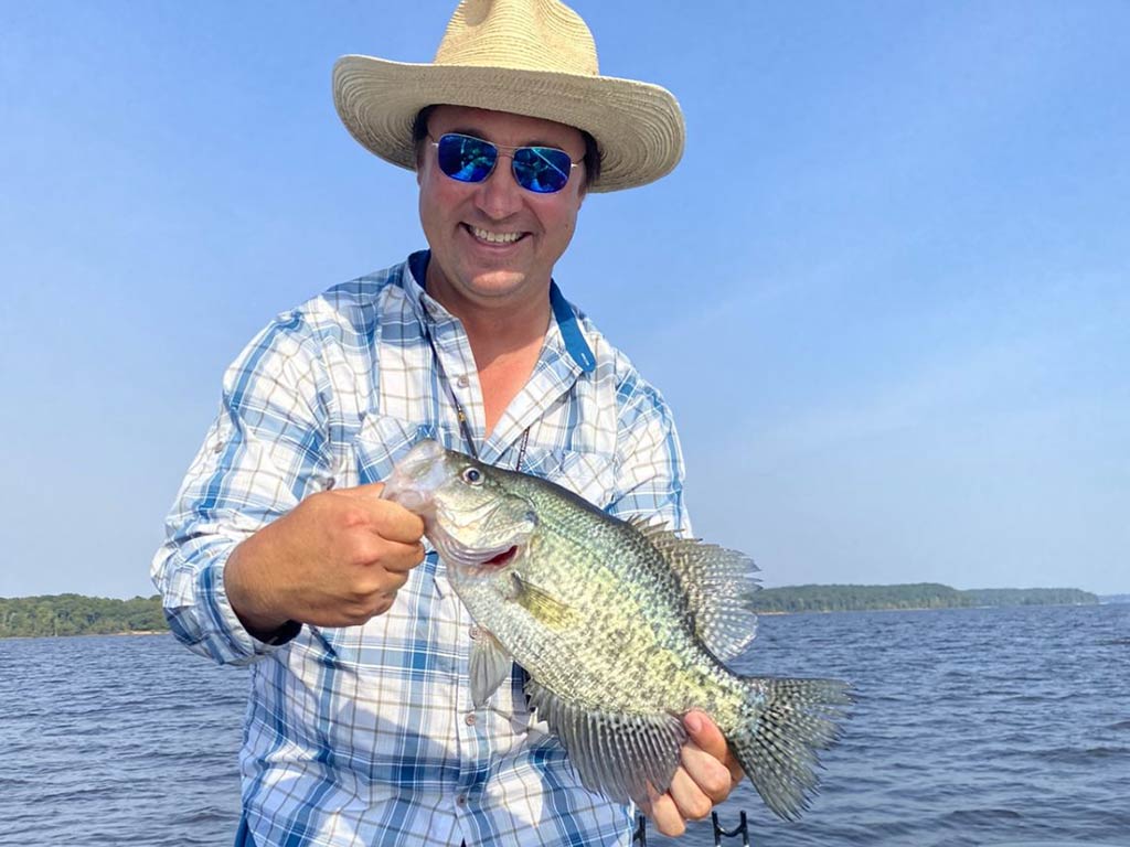 An angler in a straw hat standing on a boat on a lake in Tennessee holding a sizeable Crappie on a clear day with the water visible behind him