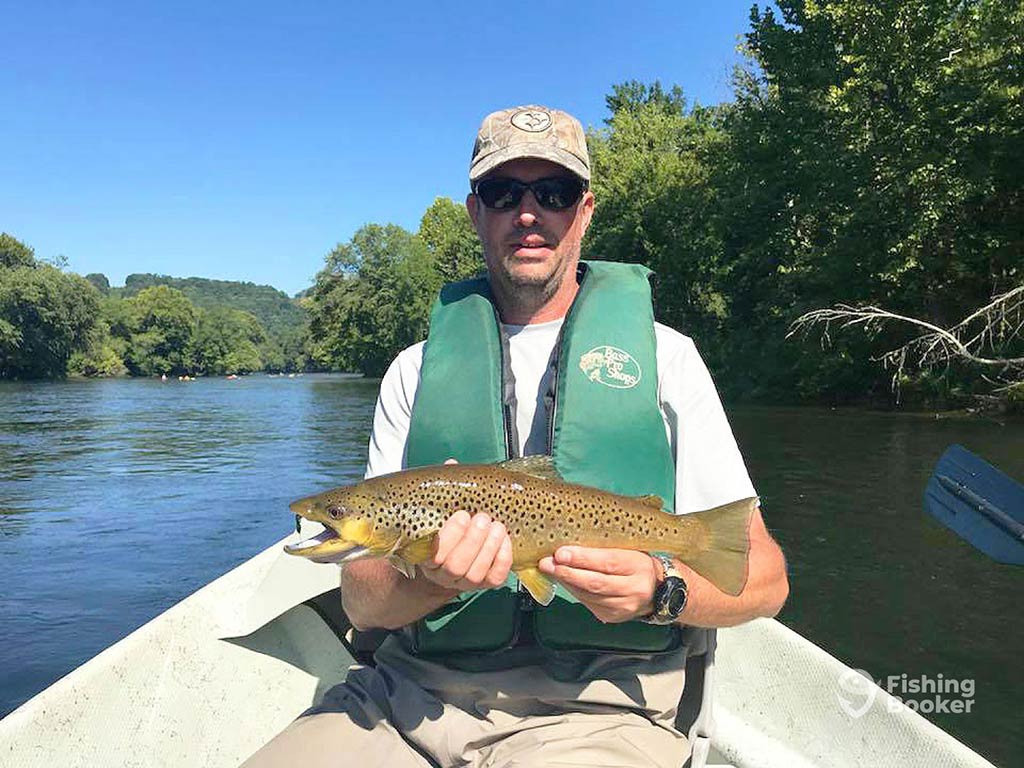 An angler wearing a personal floating device, a baseball cap, and sunglasses sitting on a boat on a river and holding a Trout on a clear day