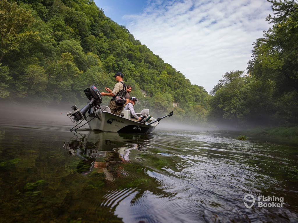 A view across the water towards an aluminum fishing boat with anglers fly fishing from it on a river in Tennessee, with the river surrounded by lush green trees