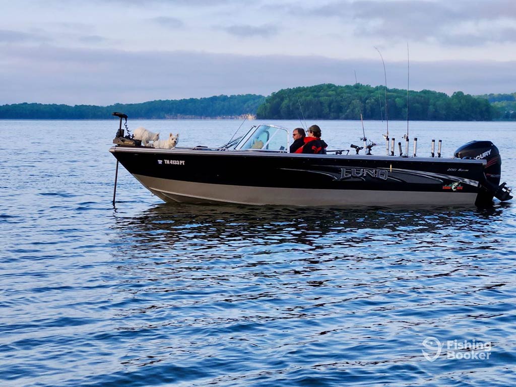 A view across the water towards a charter fishing boat on a lake in Nashville, TN near sunset on a cloudy day with two anglers aboard the boat and trolling gear on the back of the boat