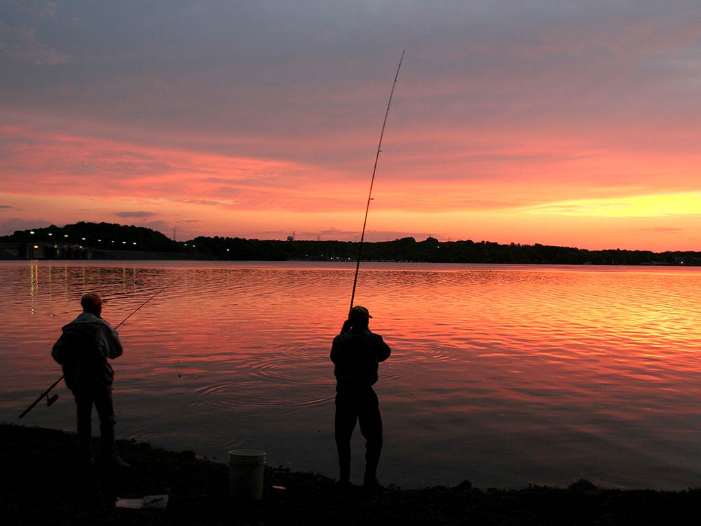 A silhouette of two men fishing at dawn on a lake in Nashville, TN, with the sun rising creating an orange hue in the distance that's also reflected on the water