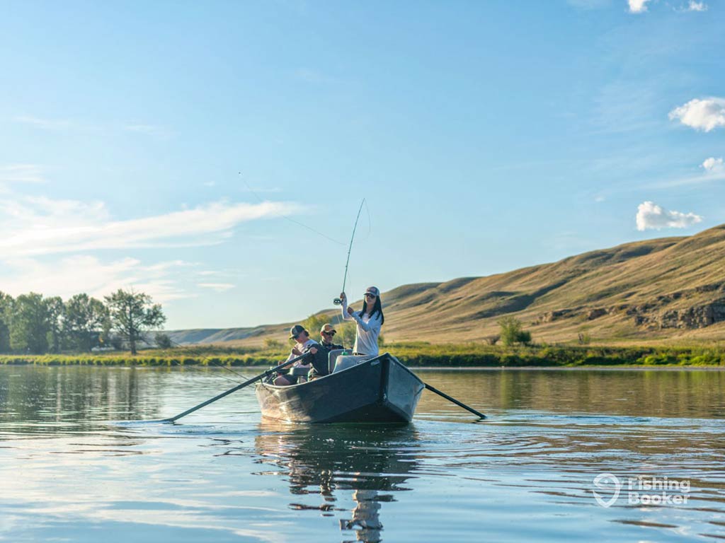 A group of anglers on a rowing boat on a clear lake on a sunny day in Alberta, with the woman at the front of the boat casting a fly rod