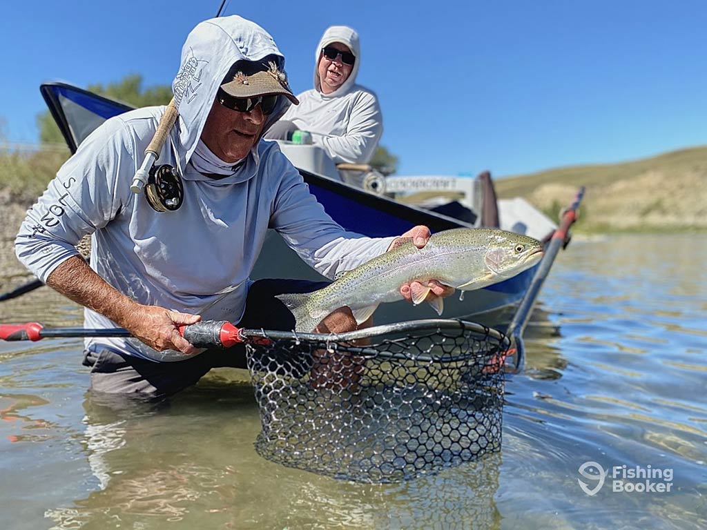 A man wading up to his waist in a river in Alberta on a clear day, holding a trout in one hand and a fishing net in the other with another man guiding a boat behind him