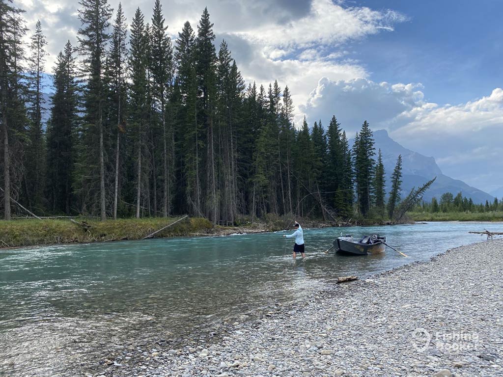 An angler wades in a gravel bed river lined with tall evergreen trees with mountains in the background, as he casts a fly with his a row boat behind him