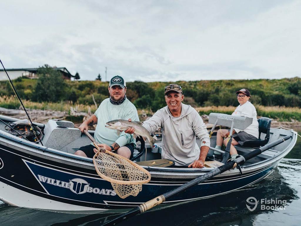 A group of three anglers aboard a light sportfishing boat on a river in Alberta on a cloudy day with two men leaning over the side of the boat in the foreground as one holds a fishing net and the other a Trout