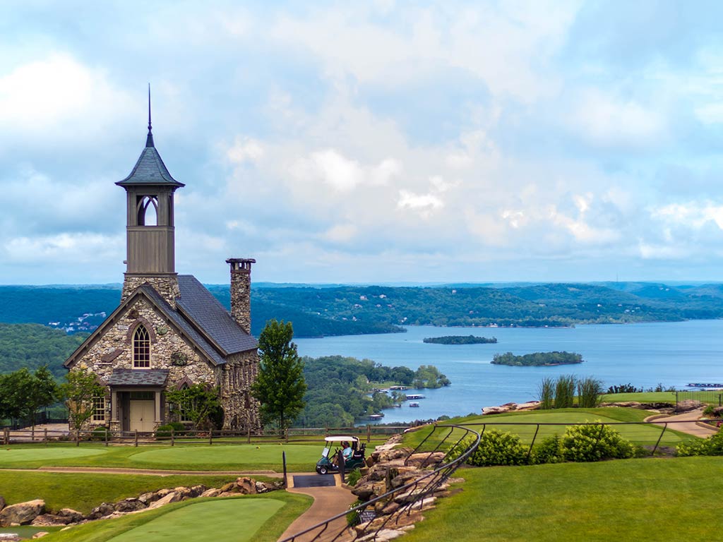 A view of a church on a hill overlooking a lake in Branson, MO on a cloudy day, with a few green islands visible in the lake in the distance