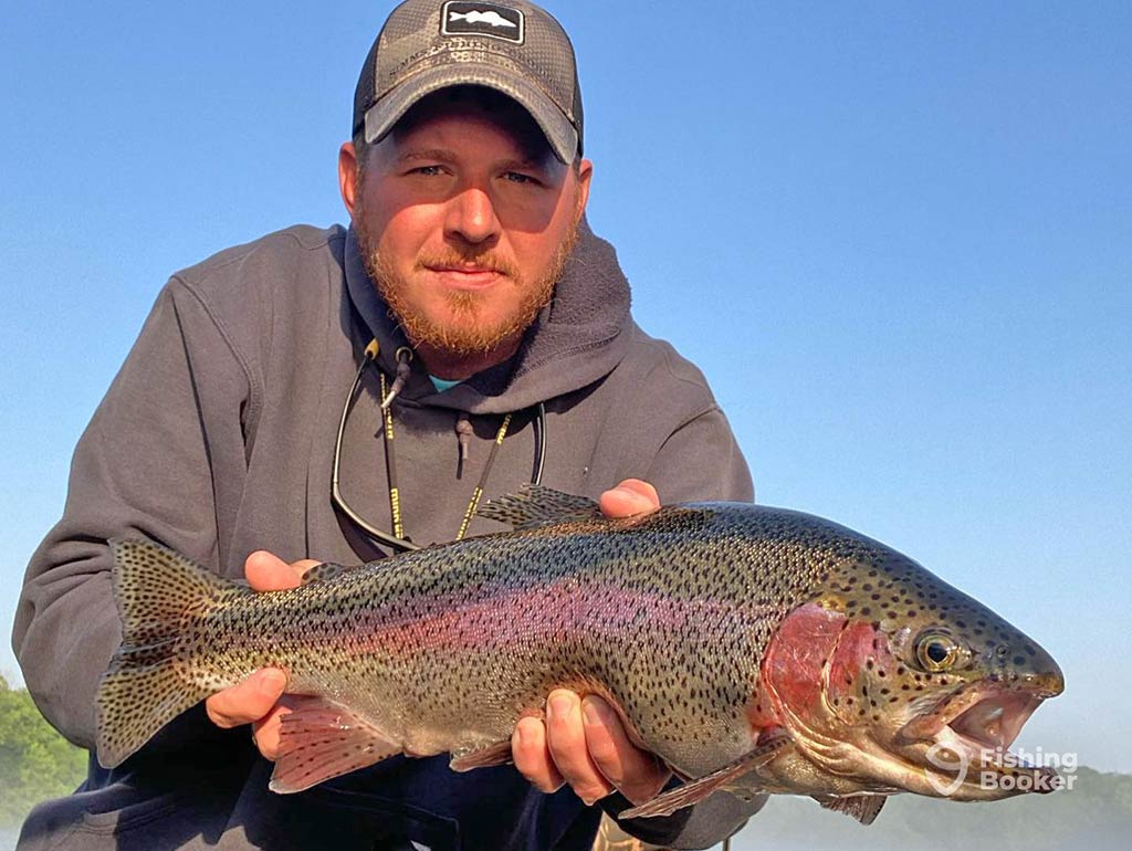 A closeup of a man in a baseball cap and hoodie holding a Trout to the camera on a clear day, with the water and blue skies visible behind him