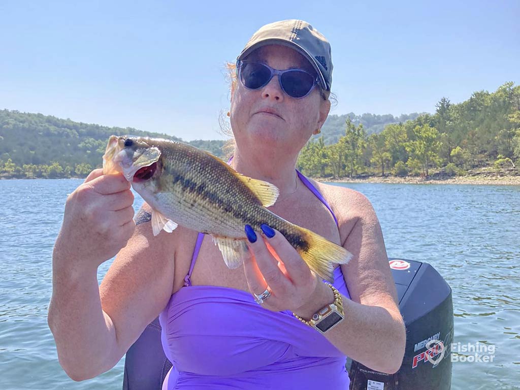 A woman in a baseball cap and sunglasses holding a small Largemouth Bass aboard a fishing boat on a lake in Branson, MO on a sunny day
