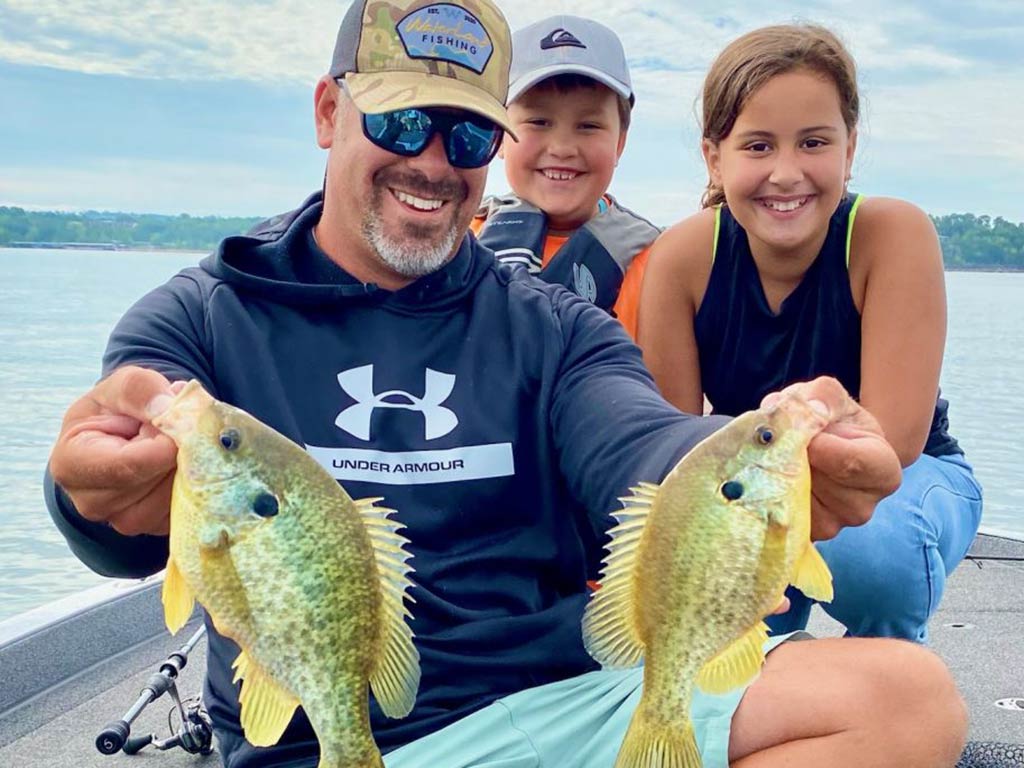 A man and two children present two Bluegill fish to the camera on a cloudy day aboard a boat in Branson, MO