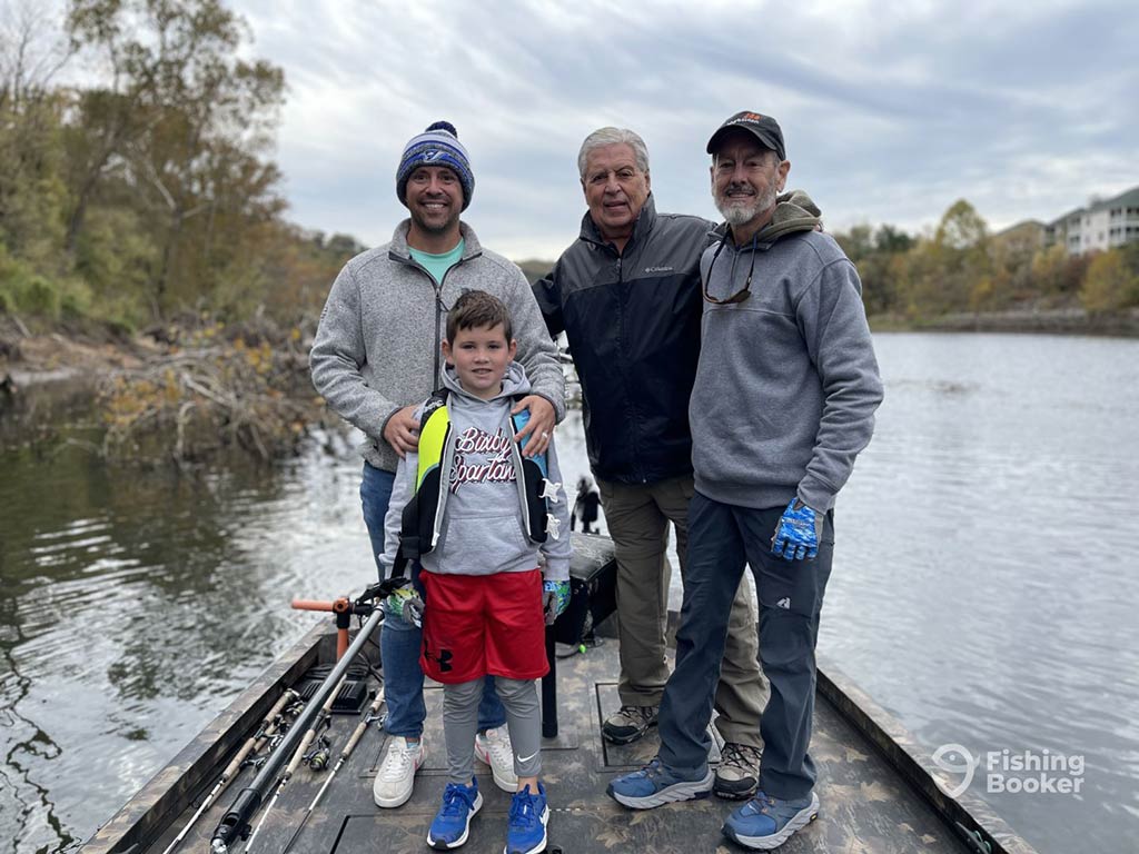 A family of three men and one boy aboard a fishing boat in Branson, MO, on a cloudy day with a number of rods visible on the left of the boat and trees and a calm body of water visible behind them