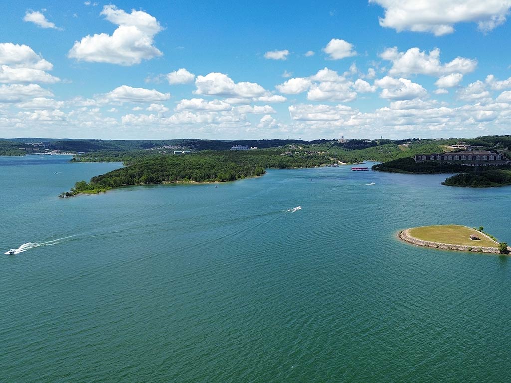 An aerial view of Table Rock Lake on a sunny day, with a boat visible on the left of the image, an island on the left, and a few green peninsulas sticking out into the lake in the distance