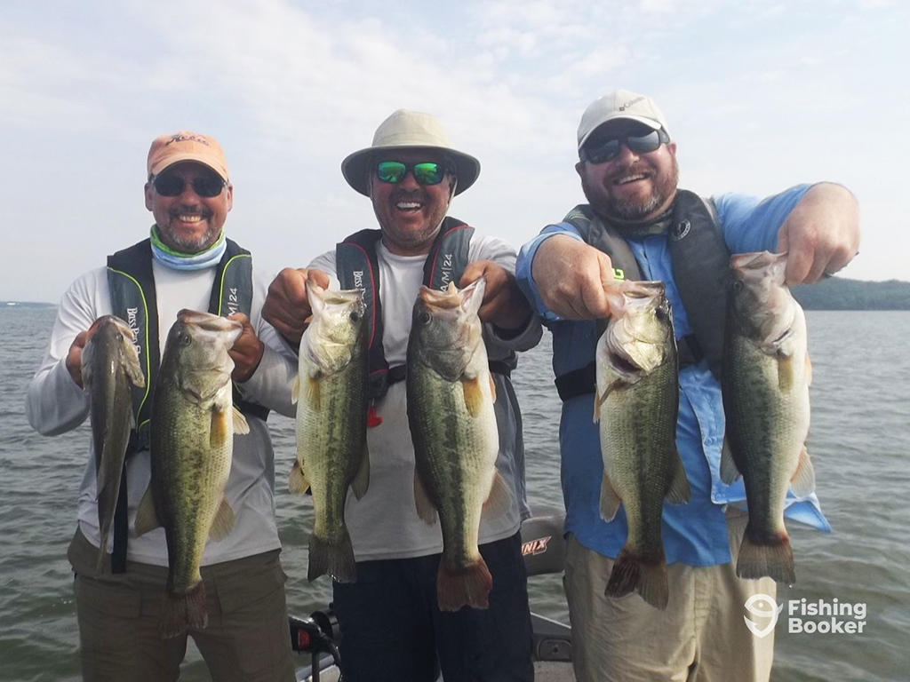 Three men in sunglasses and different hats stand at the front of a small Bass boat on a lake, each holding two Largemouth Bass