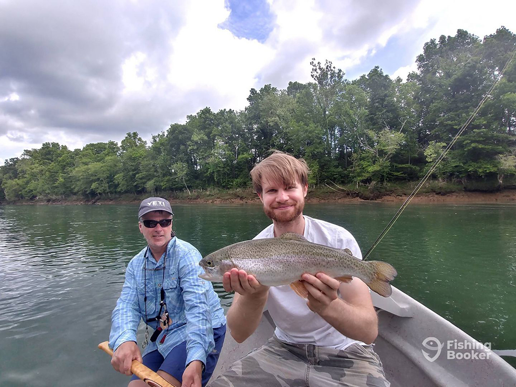 A man holds a Rainbow Trout while sitting on a rowboat on the Cumberland River in Kentucky with a fishing guide sitting behind him holding the oar of the boat