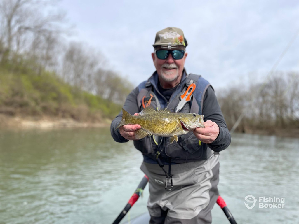 A man holds a Smallmouth Bass while standing on a small boat on a river with bare wintery trees visible on the banks around him