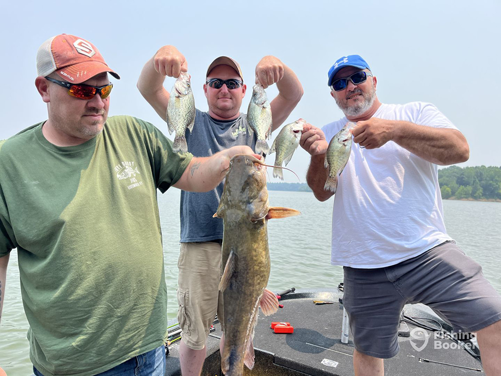 Three men stand on a small boat on a lake smiling and holding up several Crappies and a Catfish on a sunny day in Kentucky