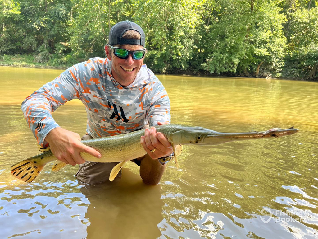 A smiling man stands knee-deep in a muddy river in the summer, holding a Longnose Gar just above the surface of the water with green trees visible behind him