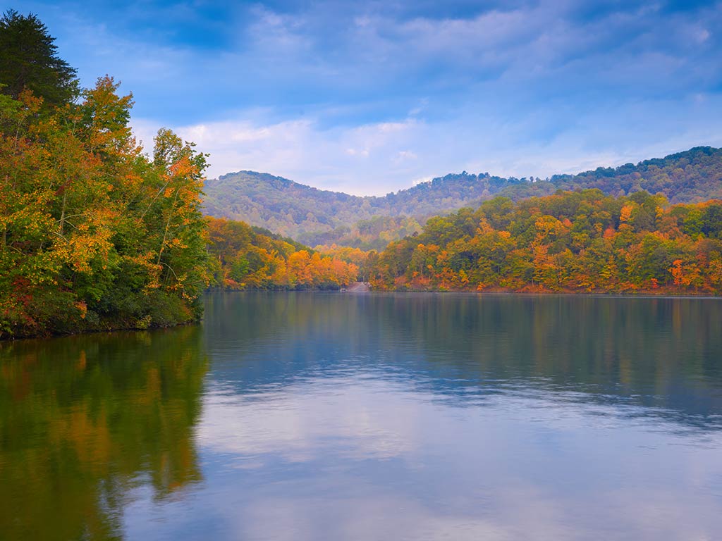 A view across a calm lake towards autumnal shorelines and hills on a day with sunny intervals