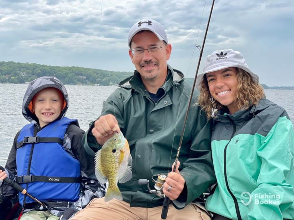 A family of three sitting on a fishing boat while wearing rain-protective gear, as the man in the middle holds a fishing rod and a Bluegill fish