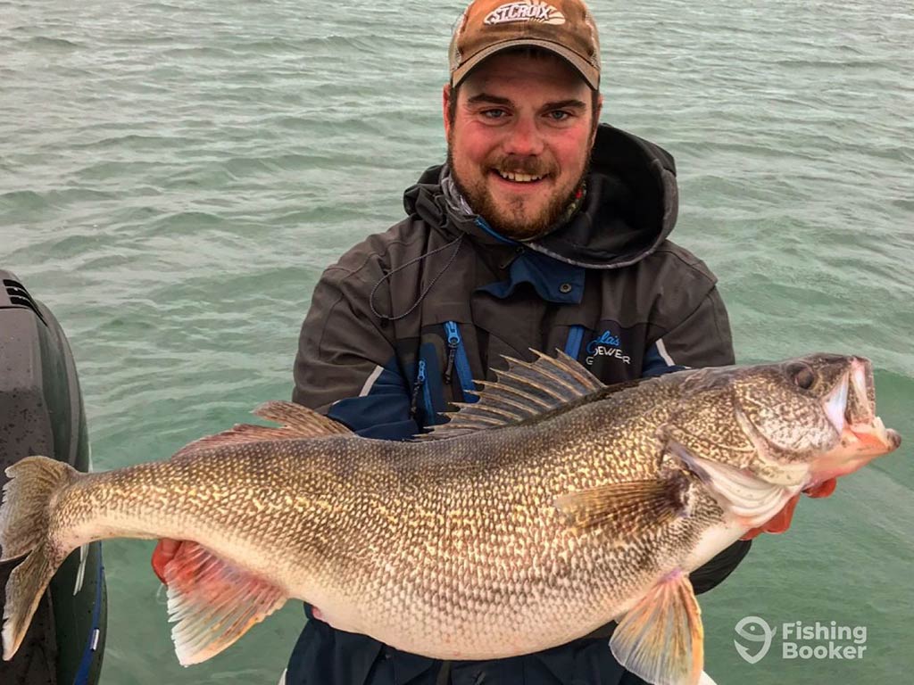 A man in a baseball cap and waterproof jacket holding a large Walleye aboard a boat on a cloudy day with murky waters visible behind him