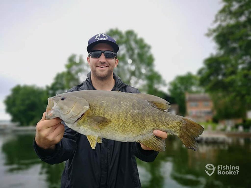 A man in a baseball cap and sunglasses holding a Smallmouth Bass next to some water on a cloudy day in Canada, with some trees visible behind him across the water