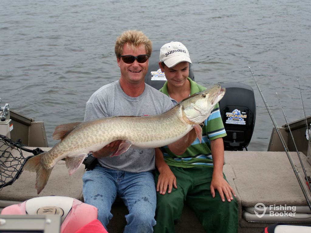 A man and a woman sitting on the deck of a fishing boat in Quebec, holding a large Northern Pike across their chest with the boat's engine and brown waters visible behind them on a cloudy day