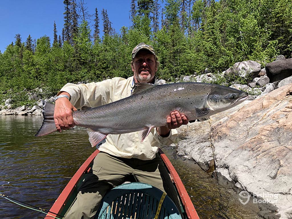 A an in a baseball cap sitting at the front of a narrow fishing boat and holding a large Salmon with a rocky shoreline and some trees visible behind him on a clear time