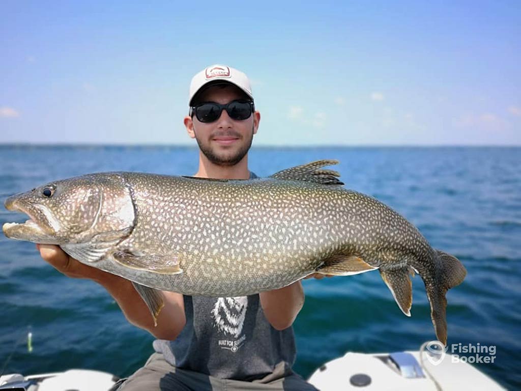 A man in a baseball cap and sunglasses holding a large Lake Trout aboard a fishing charter on a vast lake on a sunny day, with the open waters visible behind him