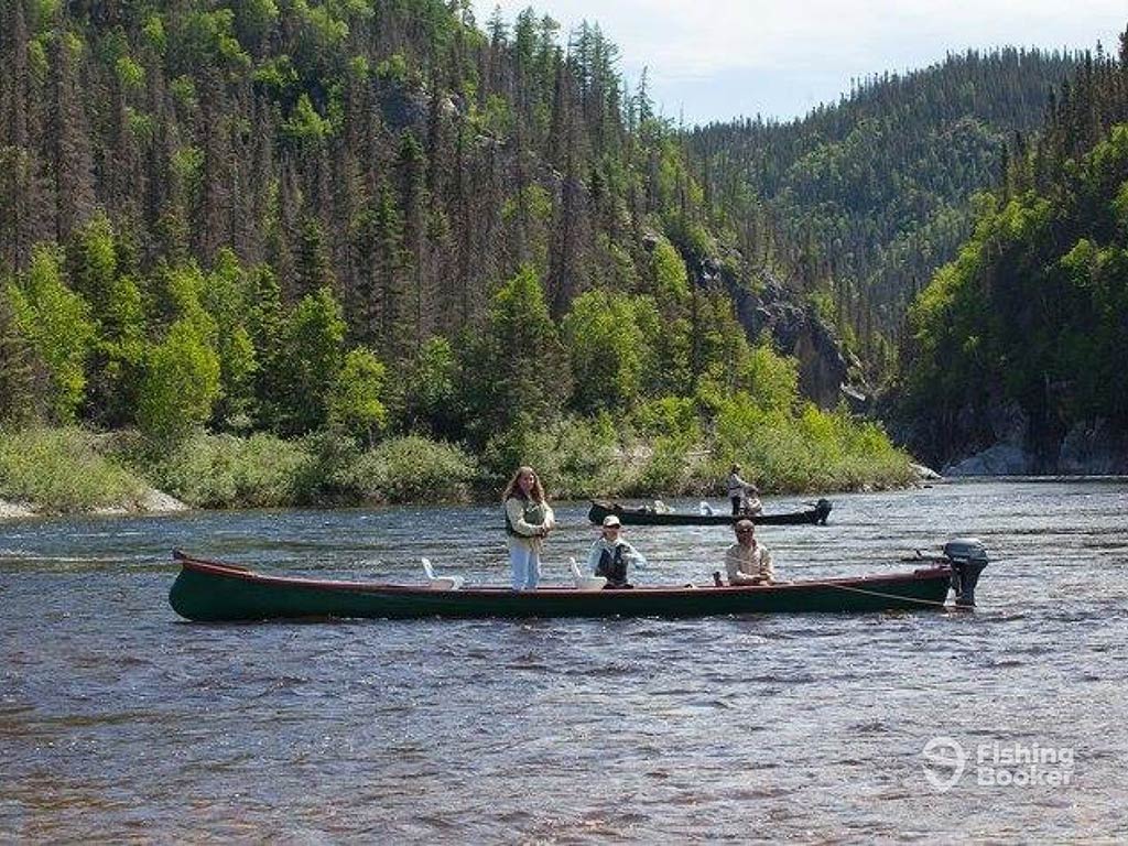 A view across the water on a river in Québec towards a long boat with three anglers enjoying a charter fishing trip on a sunny day, with trees and another boat visible behind them