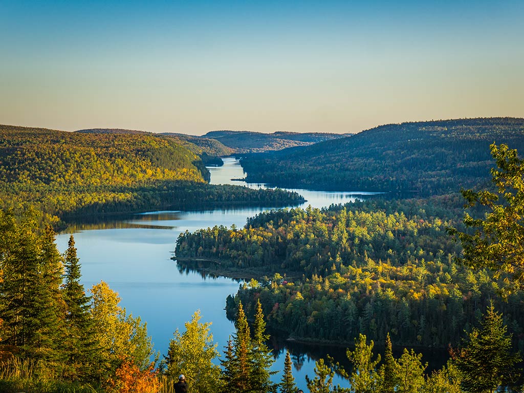 A view from a hill towards a winding lake in the Maurice National Park, Québec on a clear day, with green trees visible on either side of the blue waterway