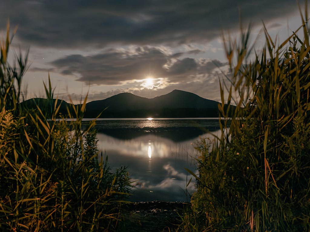 A view across the reeds towards the Lac des Deux Montagnes in Québec at sunset on a clear day, with the sun setting between the peaks of the two mountains in the distance