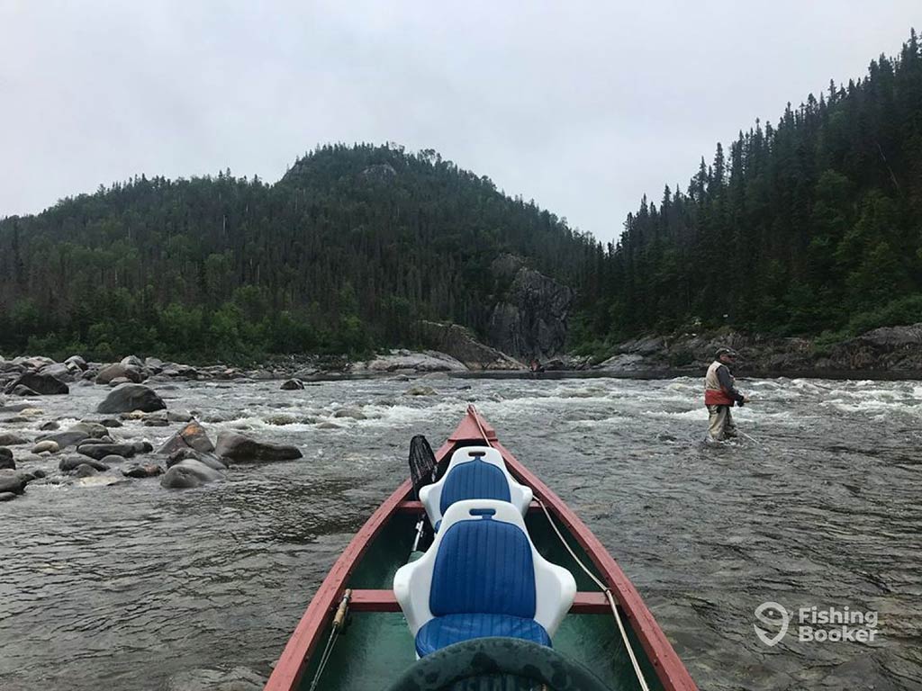 A view across the front of a narrow boat towards a man wading and fishing in a rocky river in Canada on a cloudy day, with a tree-covered hill visible in the distance