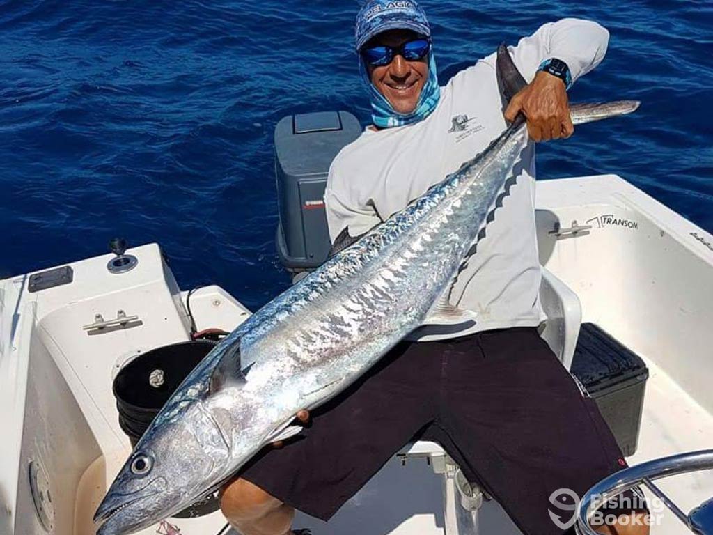 A man sitting and leaning backwards on the deck of a fishing boat on a sunny day as he holds a large Kingfish across his chest
