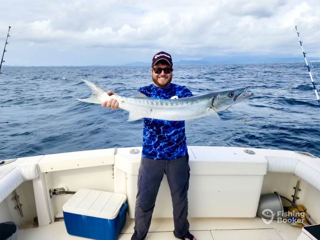 An angler in a blue shirt, sunglasses, and a baseball cap standing alone on the deck of a fishing charter on a cloudy day and holding a sizeable Barracuda