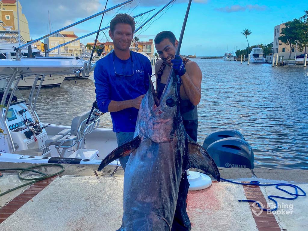 Two men standing on a dock in Puerto Rico after a successful fishing trip, holding a large Swordfish by the bill, with the water visible behind them on a clear day