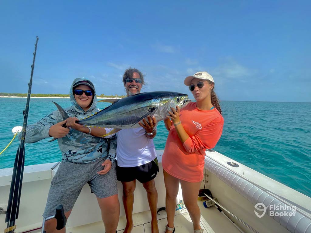 Two men and a woman standing aboard the deck of a fishing charter in San Juan, Puerto Rico holding a Yellowfin Tuna and posing with it, with crystal clear waters behind them on a clear day