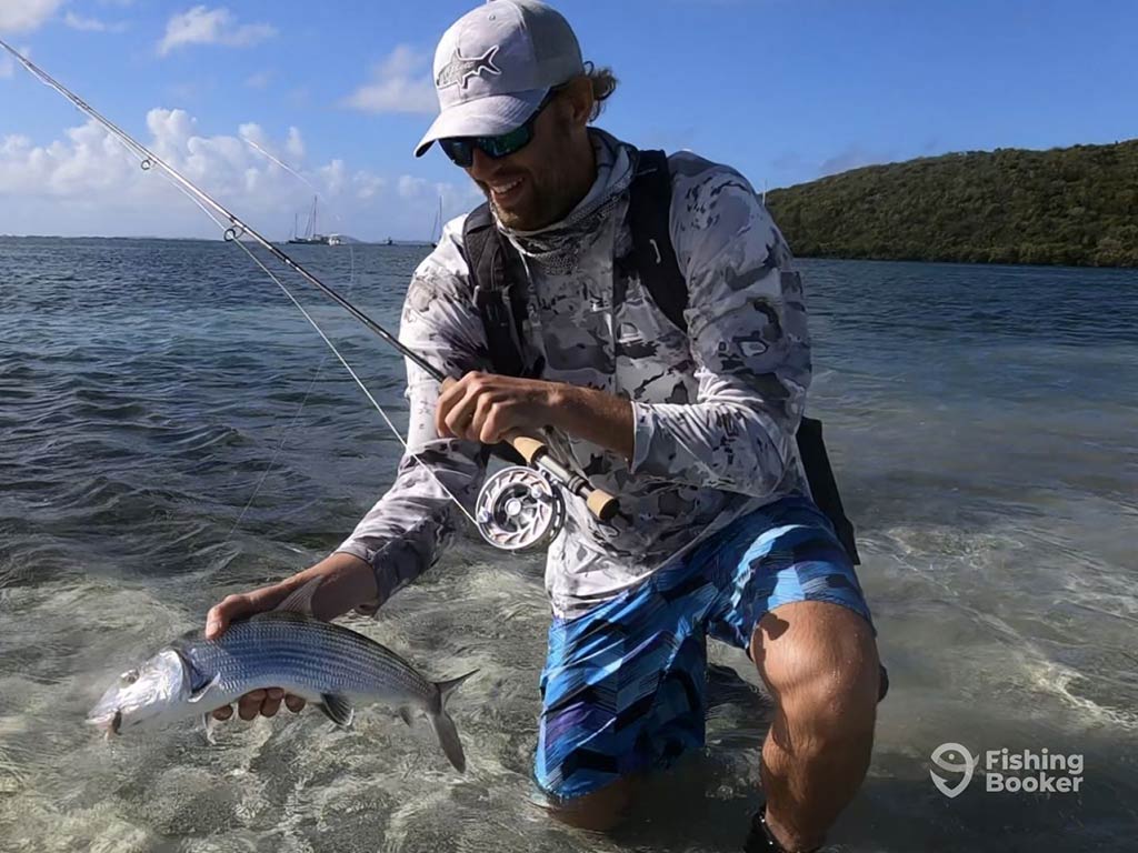 A man in a baseball cap kneeling in the shallow inshore waters of San Juan, Puerto Rico on a clear day and holding a fly fishing rod in one hand and a Bonefish on the other