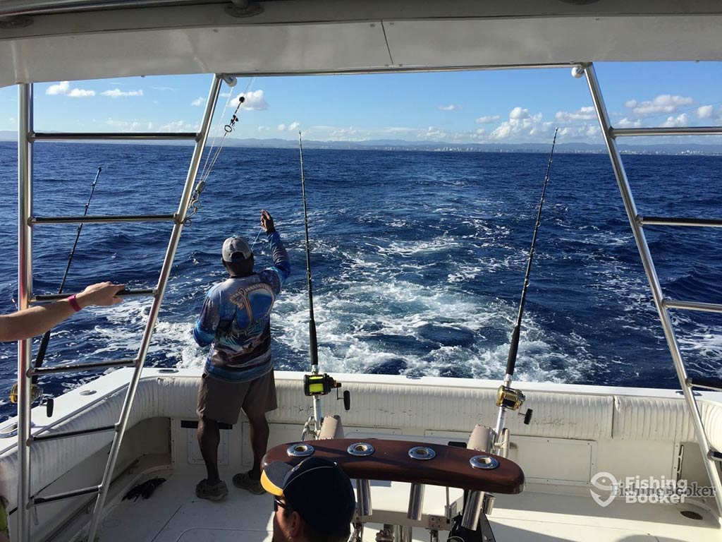 A view out of the back of a deep sea fishing charter in San Juan, Puerto Rico, with a deckhand standing on the deck tending to one of the two trolling lines, with the wake of the boat visible in the water
