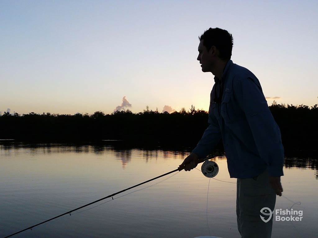 A silhouette of a man fly fishing against the backdrop of the sun setting behind a shoreline in the distance, with calm waters visible around the an on a clear day