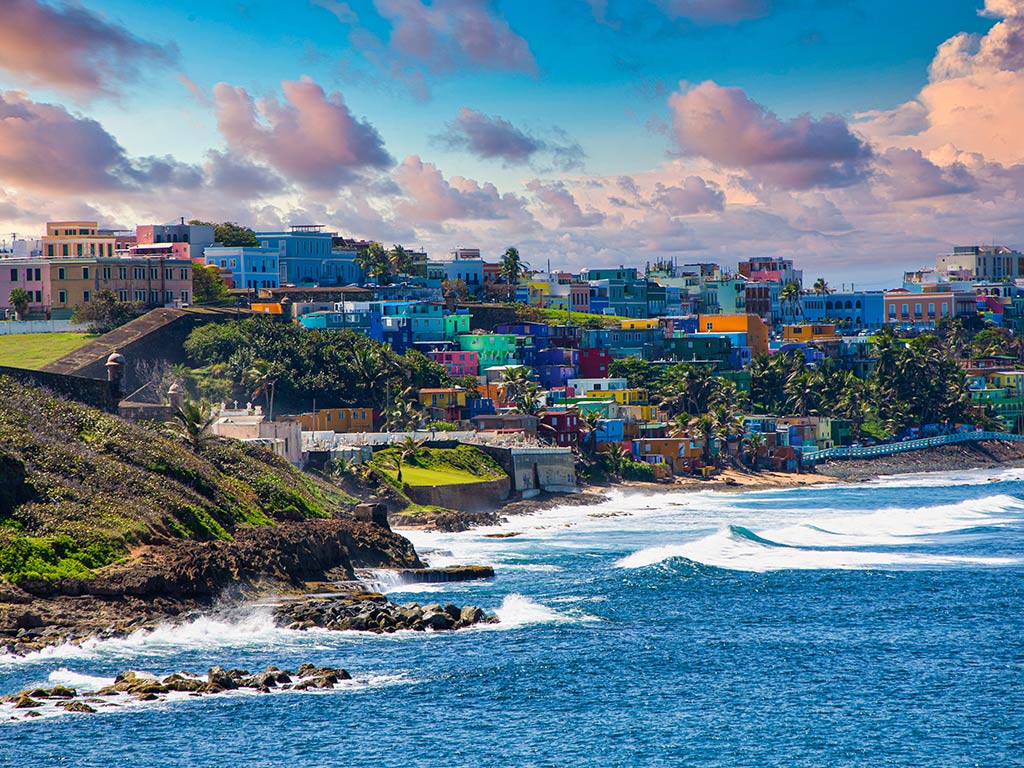 A view along the coastline of San Juan, Puerto Rico, with waves crashing into the rocky shore beneath colored houses near sunset on a clear day