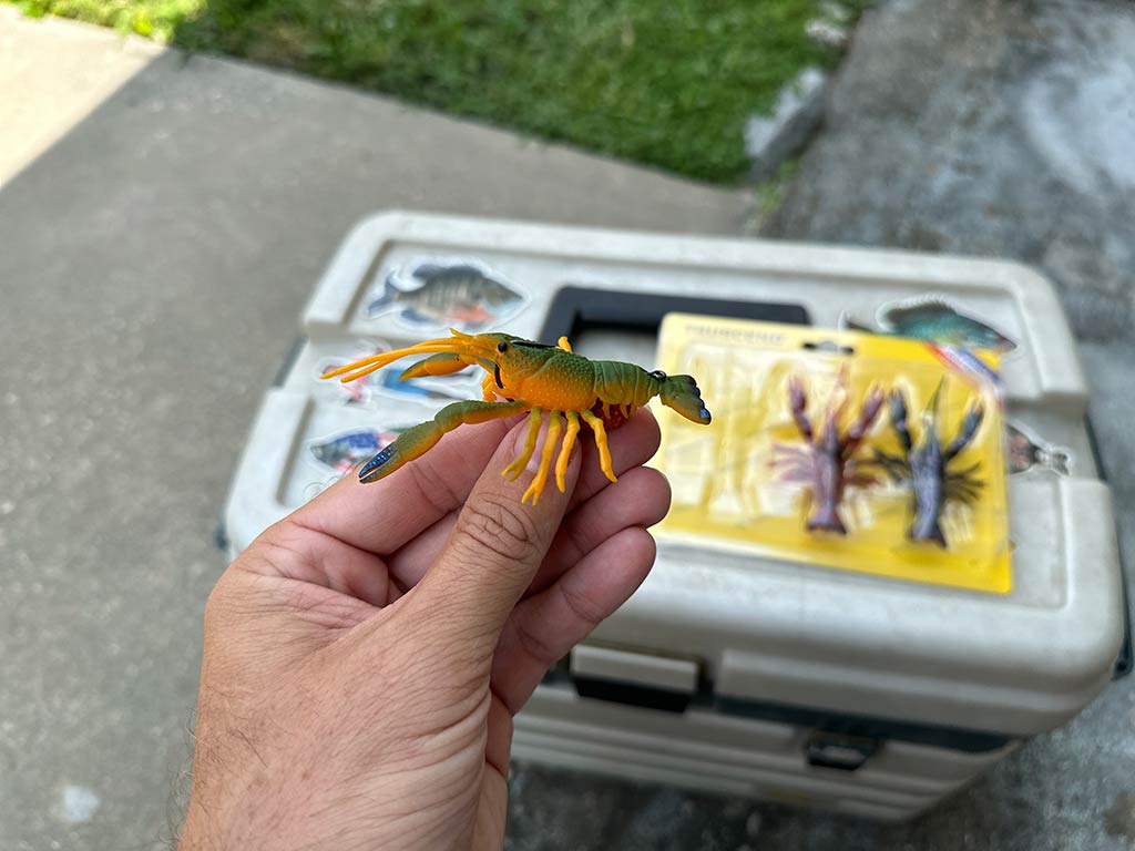 A closeup of a crawfish lure being held above a yellow-covered tackle box on dry land