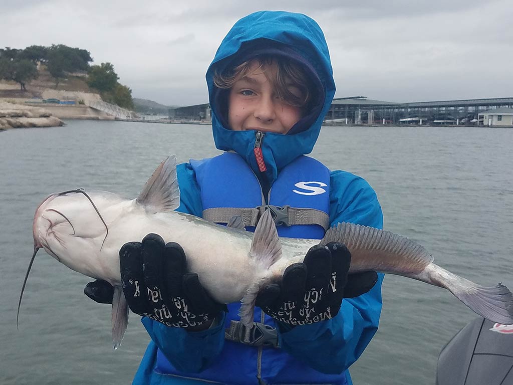 A young male angler in a blue winter jacket with the hood up, holding a Catfish caught on Lake Travis while standing on a boat with the water and grey skies behind him
