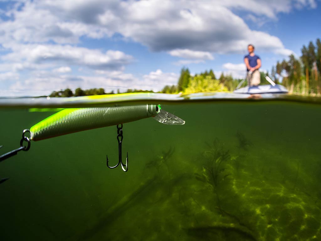 A split shot of the man fishing from the boat on the lake, with an underwater view of a jerkbait in the water