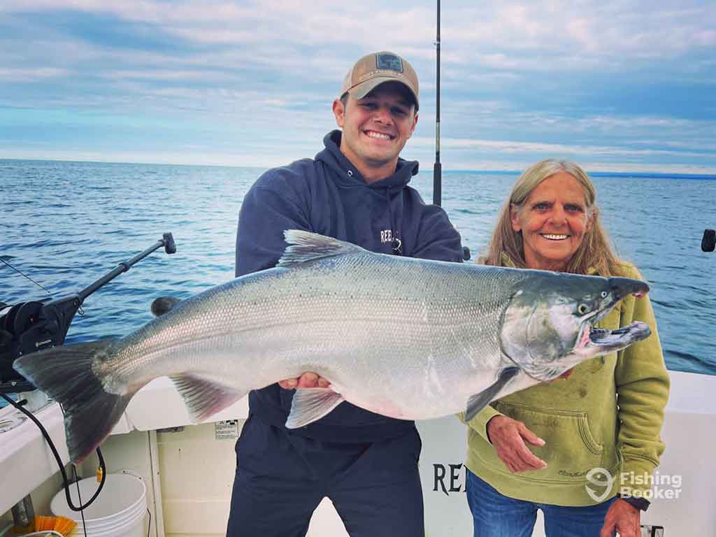 A man and a woman standing on a fishing boat in upstate New York, holding a large King Salmon between them on a cloudy day with calm waters visible behind them