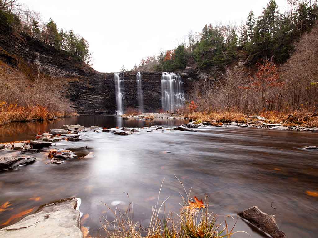 A view towards a waterfall in Salmon River, NY, with fall foliage all around on a cloudy day